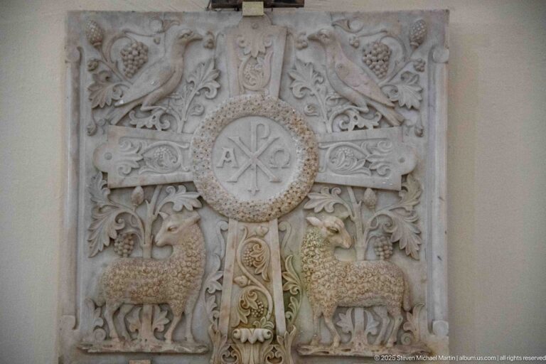 Carved stone relief with Chi-Rho medallion, broad cross, two facing lambs, vines, leaves, and fruit clusters.