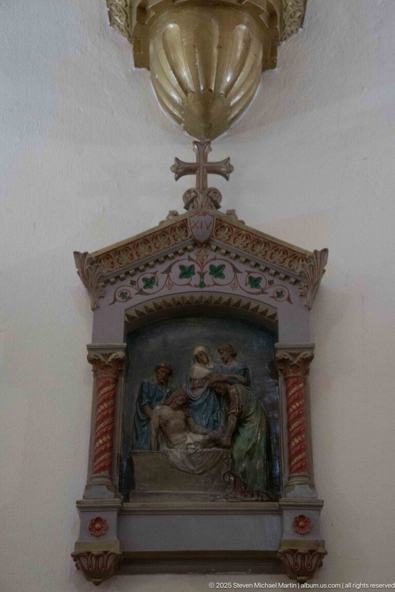 Wall-mounted devotional relief showing robed figures around a reclining figure beneath a suspended metal lamp and small architectural frame.