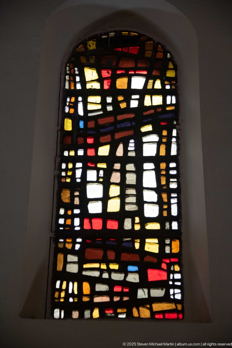 Arched abstract stained-glass window with irregular black leading and scattered amber, red, blue, white, and yellow panes.