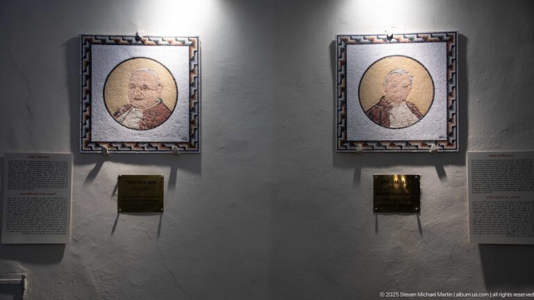 Two illuminated framed papal portraits on a white wall with small plaques and explanatory text panels beneath and beside them.