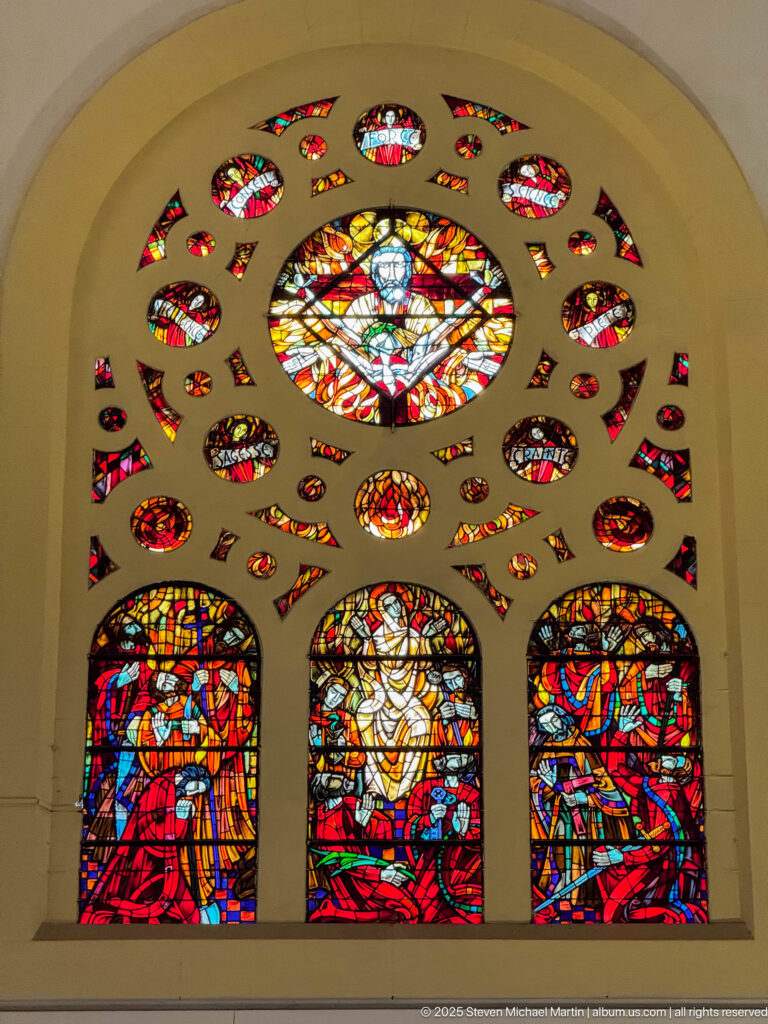 Large stained-glass ensemble with three figurative lower panels, central circular medallion, and many smaller multicolored panes above.