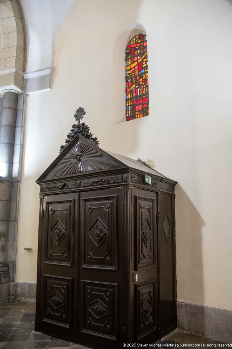 Dark wooden confessional with paneled doors, carved geometric ornament, triangular pediment, and small cross beneath a stained-glass window.