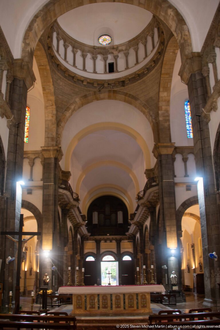 Centered nave view toward the rear entrance and organ gallery, framed by stone arches, piers, upper balustrade, altar, and small stained-glass windows.