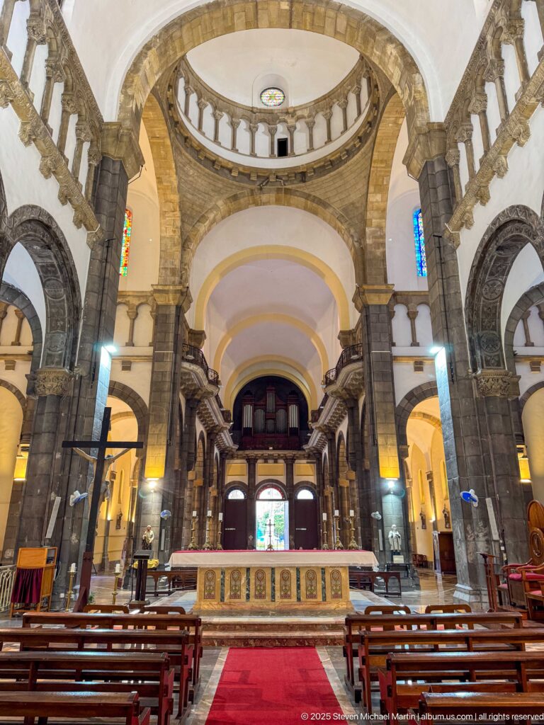 Nave interior with altar, red carpet, wooden pews, dark stone arches, piers, circular upper balustrade, and rear gallery.