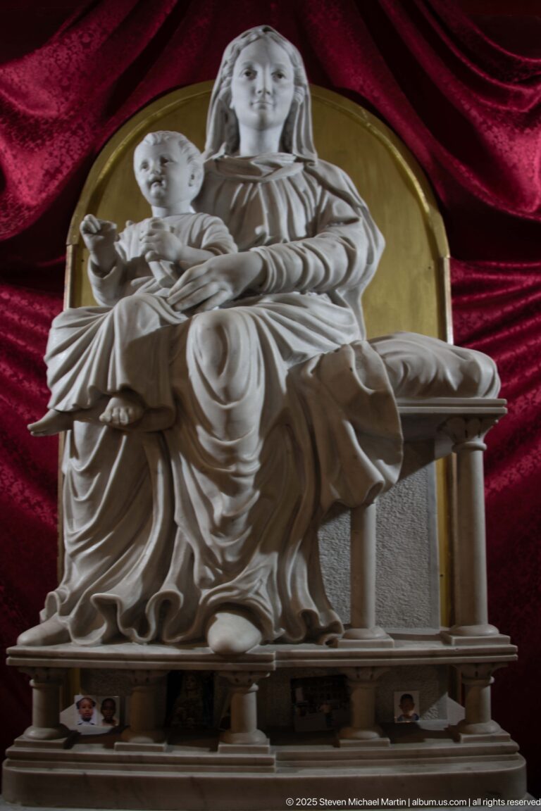 White marble Madonna and Child sculpture against deep red velvet curtains and a gold-toned circular backdrop.