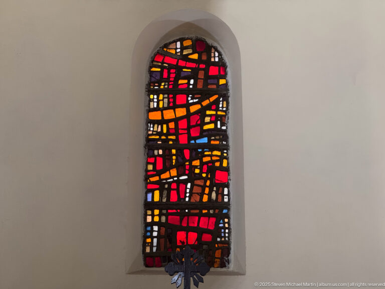 Narrow arched stained-glass window with irregular red, orange, amber, black, and blue panes set in a pale wall.