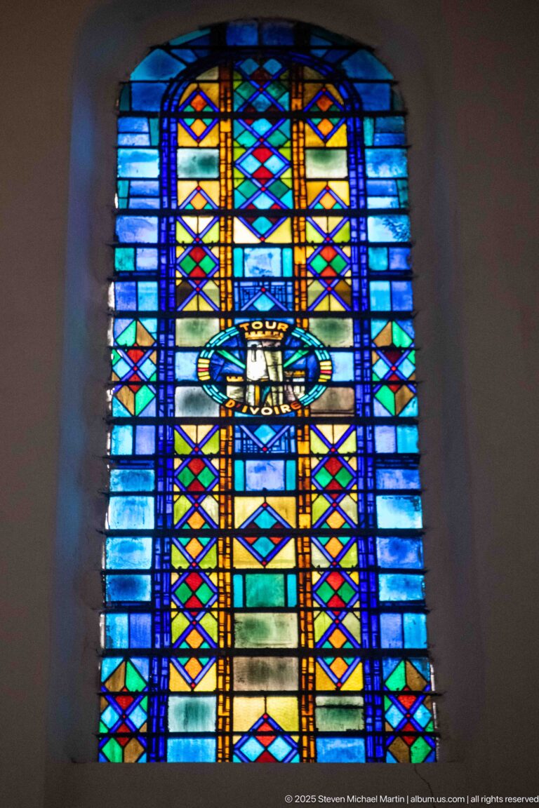 Blue-dominant stained-glass window with central oval emblem and symmetrical yellow, red, green, and orange geometric bands.
