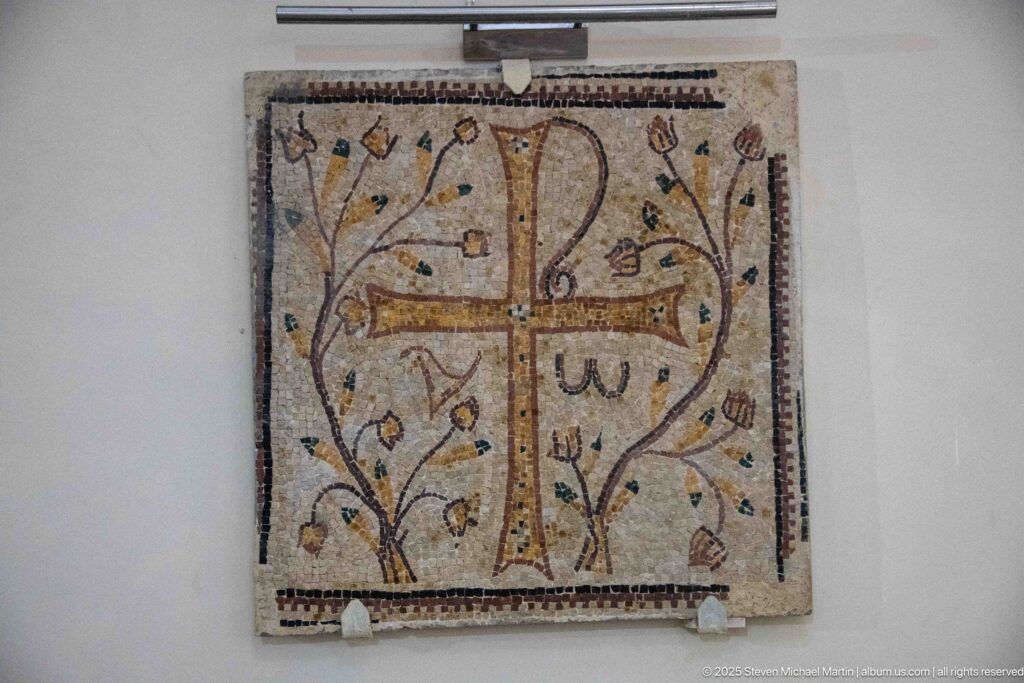 Square mosaic with jeweled cross, Alpha and Omega symbols, curling vines, small bird motifs, and patterned border.