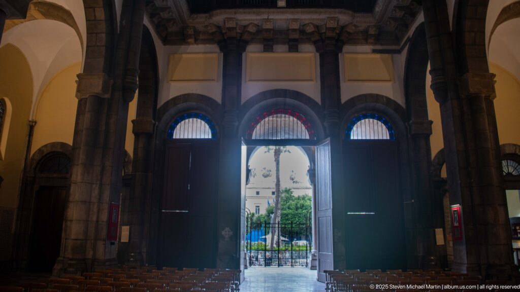 Rear nave view toward the bright main entrance, framed by dark stone piers and three arched doorways with wooden chairs in the foreground.