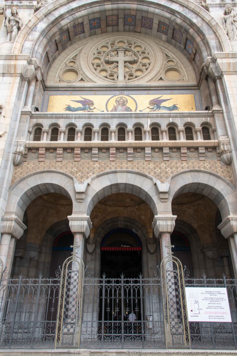 Three arched cathedral entrances with columns and iron gates beneath a gold mosaic band and circular stone tracery.