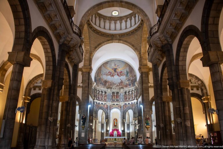 Long nave view toward the sanctuary with massive stone arches, upper balustrade, apse mural, painted saints, and altar.