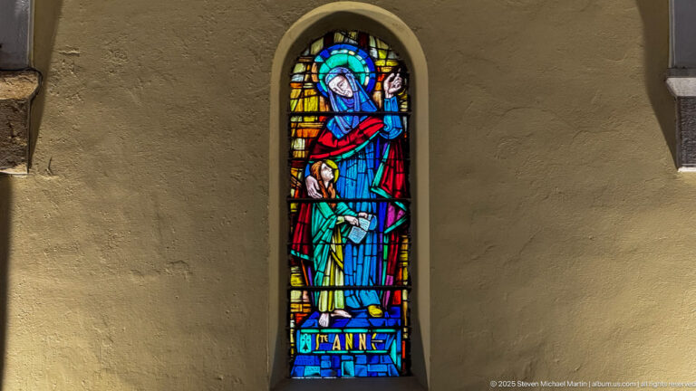 Narrow stained-glass window of Saint Anne with a smaller figure, labeled “Ste Anne” at the base in bright blue, red, and green glass.