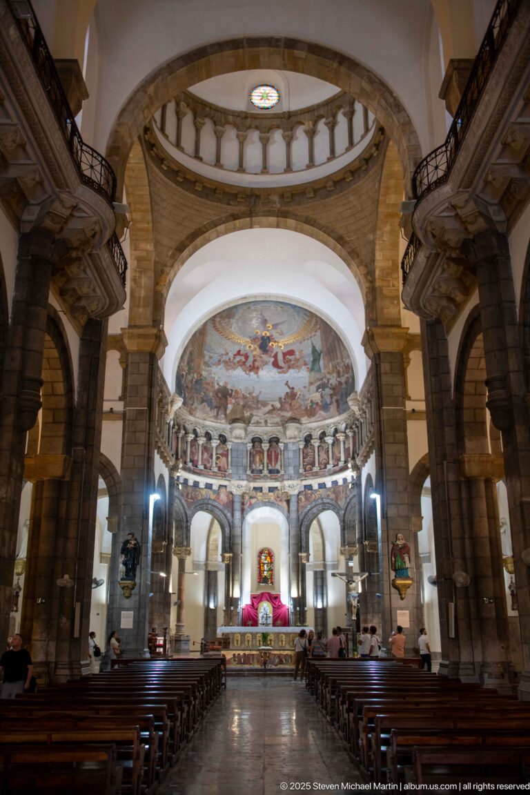 Wide nave view toward sanctuary with pews, stone piers, arches, circular balustrade, painted saints, and apse mural.