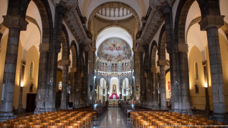 Long symmetrical aisle view toward the apse, with wooden seating, tall dark stone columns, arches, and small stained-glass windows.