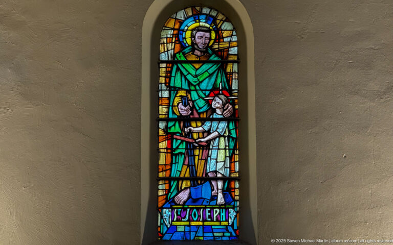 Narrow stained-glass window of Saint Joseph in green robes, labeled “St Joseph” at the base and framed by multicolored glass.