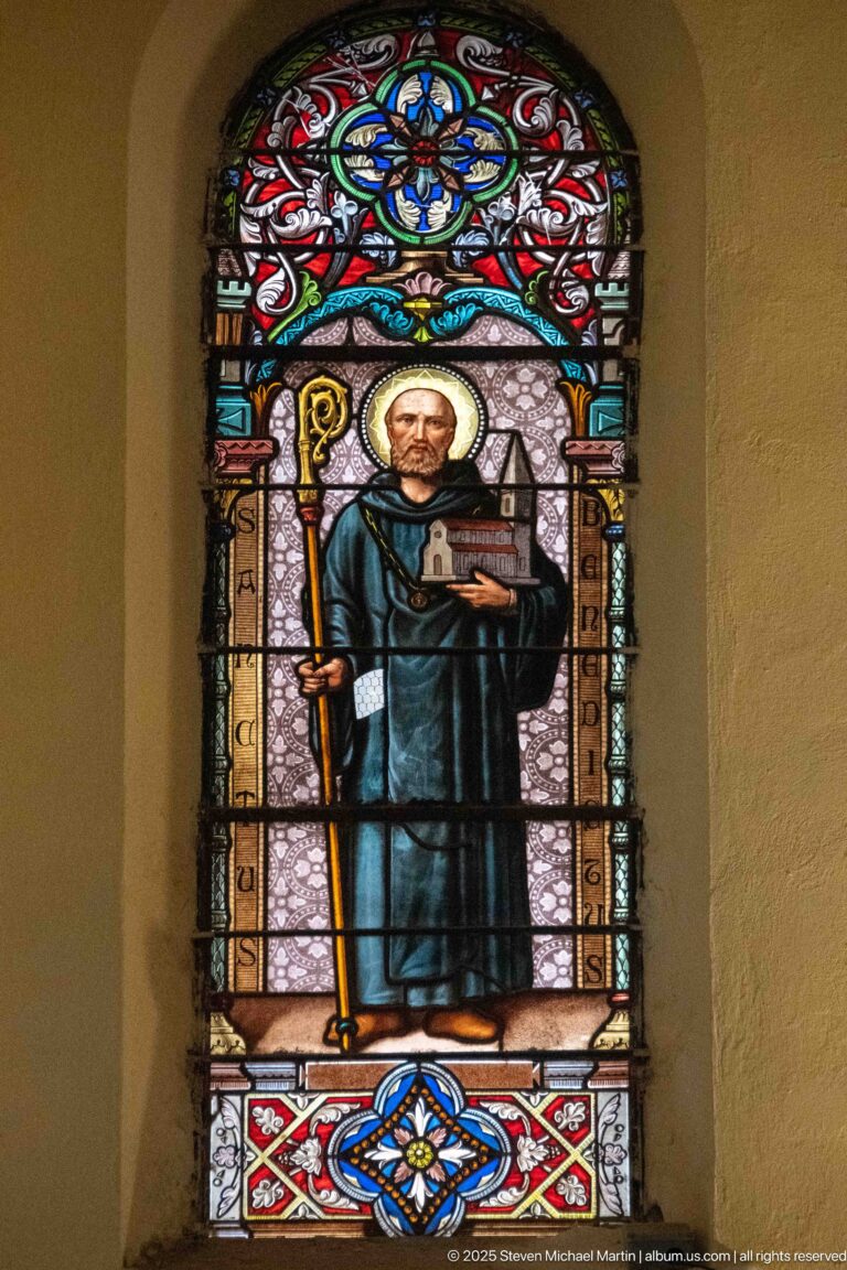 Arched stained-glass window of a haloed monastic saint in a dark habit holding a crozier and open book.