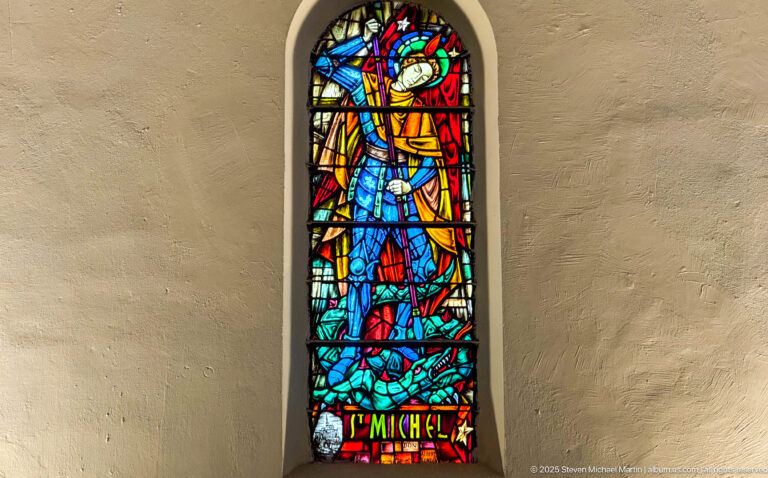 Narrow stained-glass window of Saint Michael in red, gold, and blue above a subdued figure, labeled “ST MICHEL.”