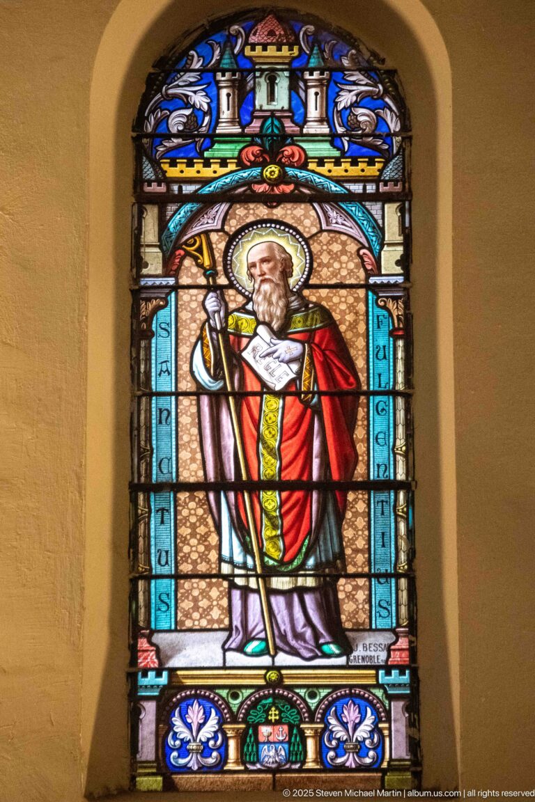 Arched stained-glass window of a haloed bearded bishop in red and gold vestments holding a chalice-like vessel.