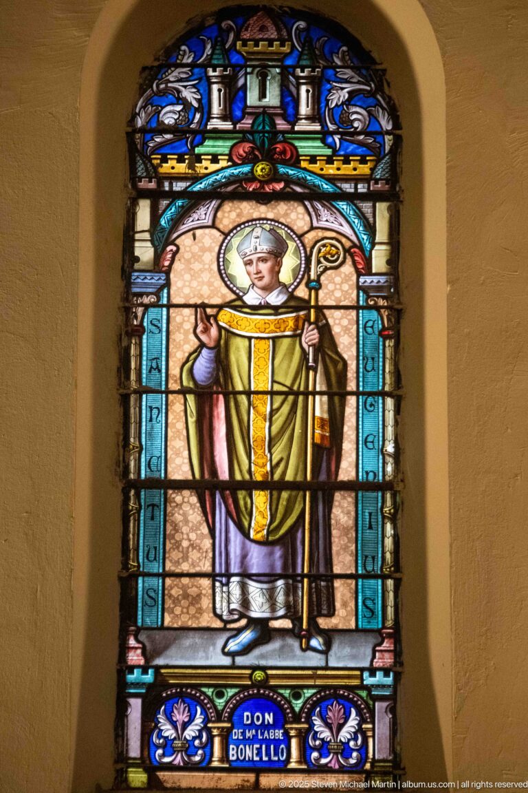 Arched stained-glass window of a haloed bishop in gold vestments holding a crozier and raising one hand in blessing.