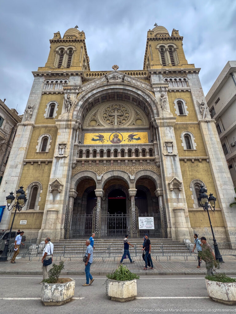 Frontal view of St. Vincent de Paul Cathedral with twin towers, monumental arch, mosaics, three portals, and pedestrians.