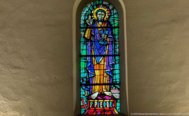 Narrow stained-glass window of Saint Peter in blue and gold robes, labeled “St Pierre” at the base and holding a key-like attribute.