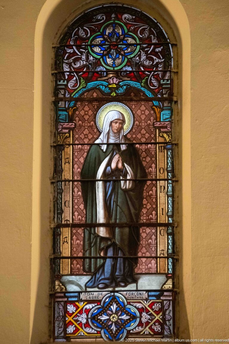 Full-length stained-glass female saint in a dark habit with crossed hands, halo, and ornate multicolored floral border.