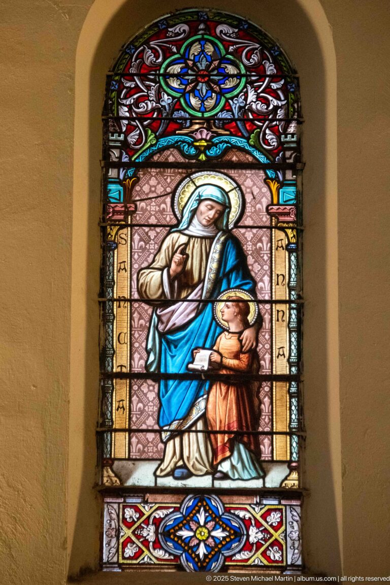 Stained-glass Saint Anne in blue and cream beside a child, framed by ornate red, blue, amber, and white glass.