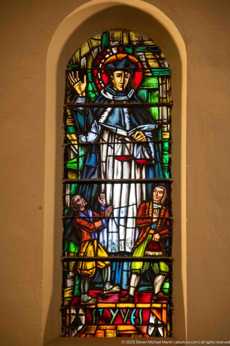 Haloed woman in pale robes above kneeling armored men in a vivid red, green, blue, and gold stained-glass scene.