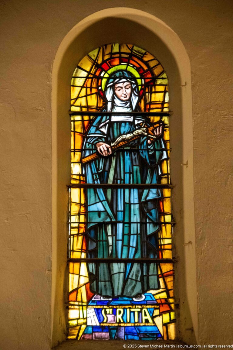 Stained-glass Saint Rita in a dark habit with halo and devotional object, labeled “ST RITA” at the base.