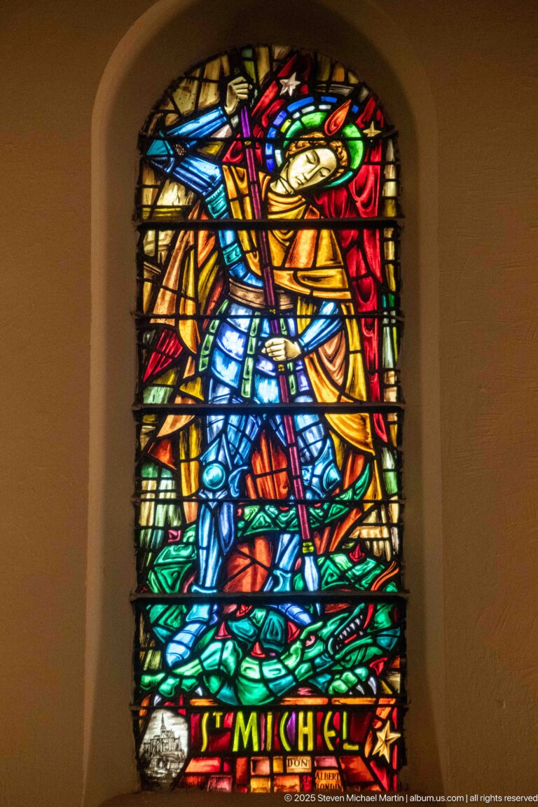 Stained-glass Saint Michael in armor standing above a defeated figure, with vivid multicolored glass and “ST MICHEL” inscription.