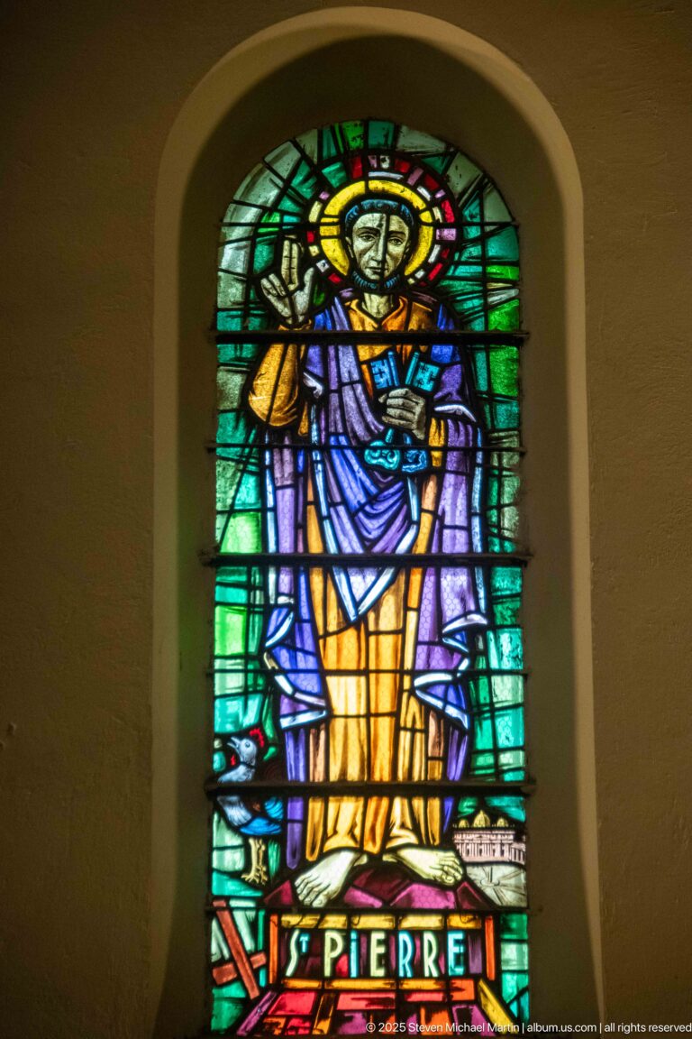 Stained-glass Saint Peter in gold, lavender, and blue robes holding a key-like attribute, labeled “ST PIERRE.”