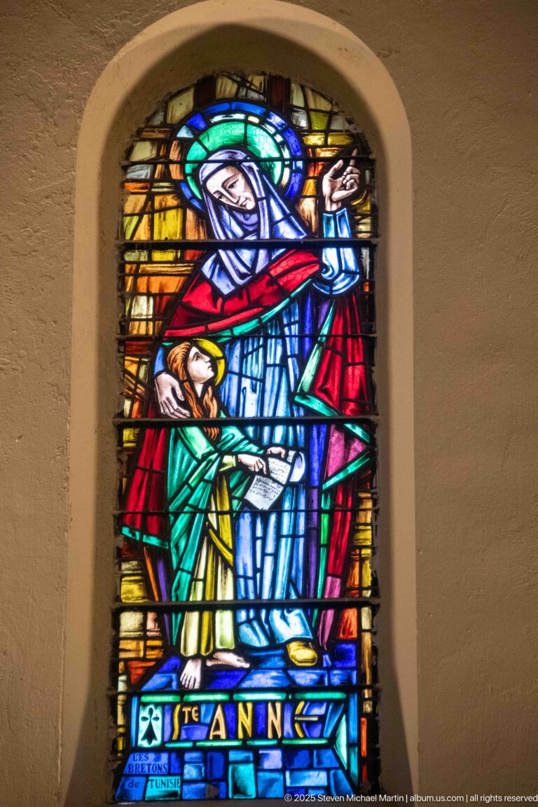 Stained-glass Saint Anne in blue and red beside a child, labeled “STE ANNE” at the base.