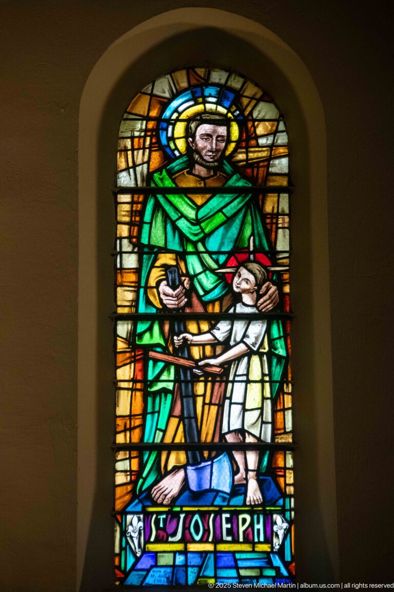 Stained-glass Saint Joseph in green robes beside the Christ Child, with halo and “ST JOSEPH” inscription.