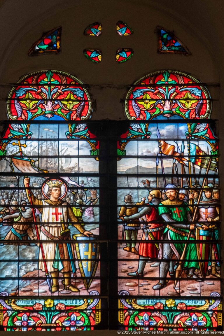 Paired stained-glass narrative scenes with robed and armored figures, military standards, weapons, and ornate floral borders.
