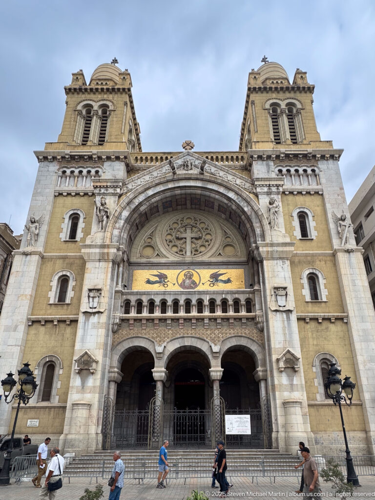 Broad frontal view of St. Vincent de Paul Cathedral, showing twin towers, central arch, rose tracery, mosaic band, and pedestrians.