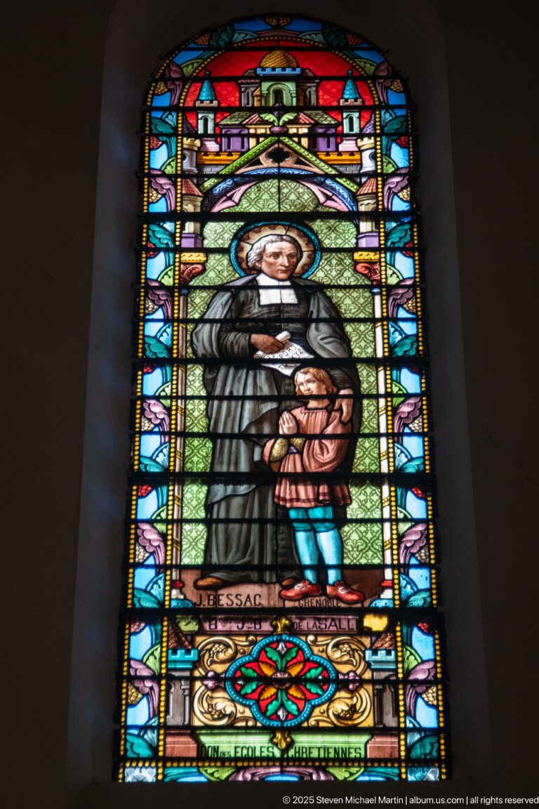 Haloed male religious figure in a dark habit standing beside a child within an ornate stained-glass architectural frame.