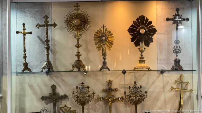Glass display case containing crosses, sunburst monstrance-like objects, ornate stands, and smaller Catholic devotional metalwork.