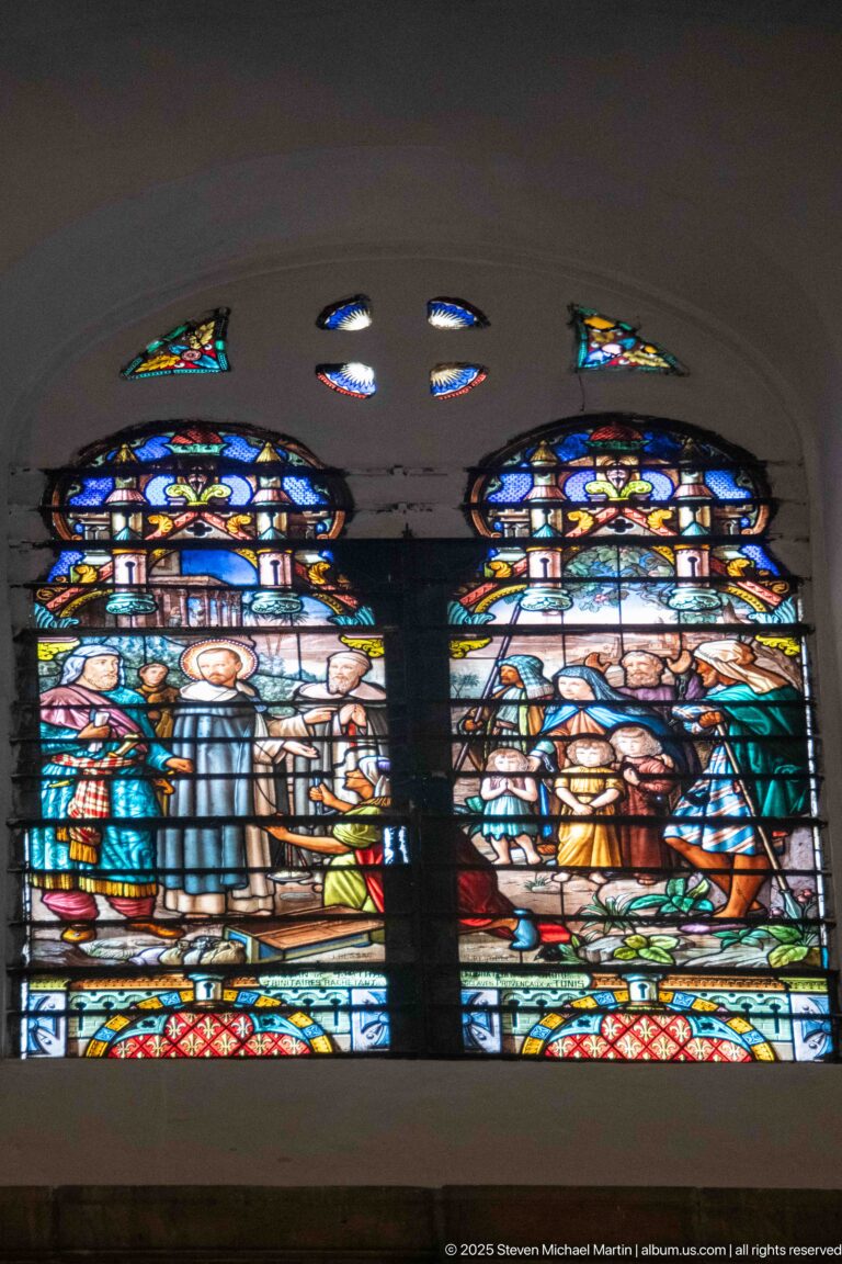 Two adjacent stained-glass religious scenes with grouped figures, decorative roundels, and vivid red, blue, green, and gold borders.