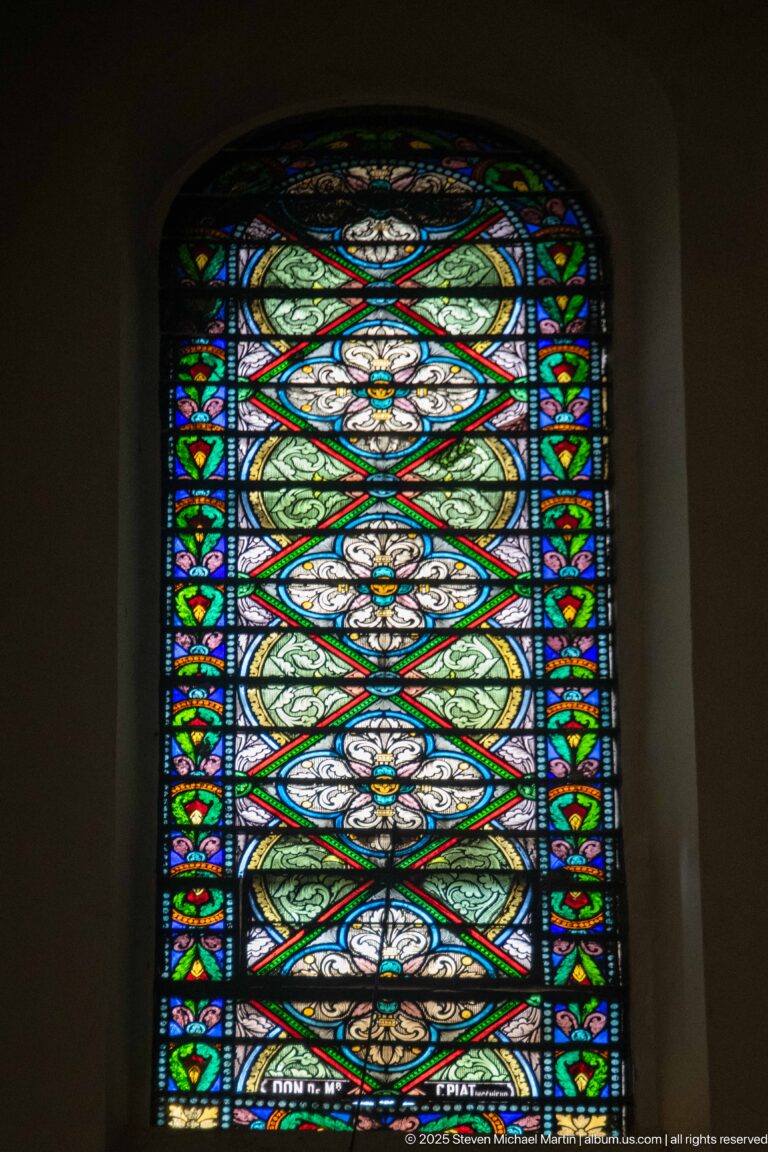 Tall ornamental stained-glass panel with pale green circular motifs, white floral centers, red diagonal bands, and blue border.
