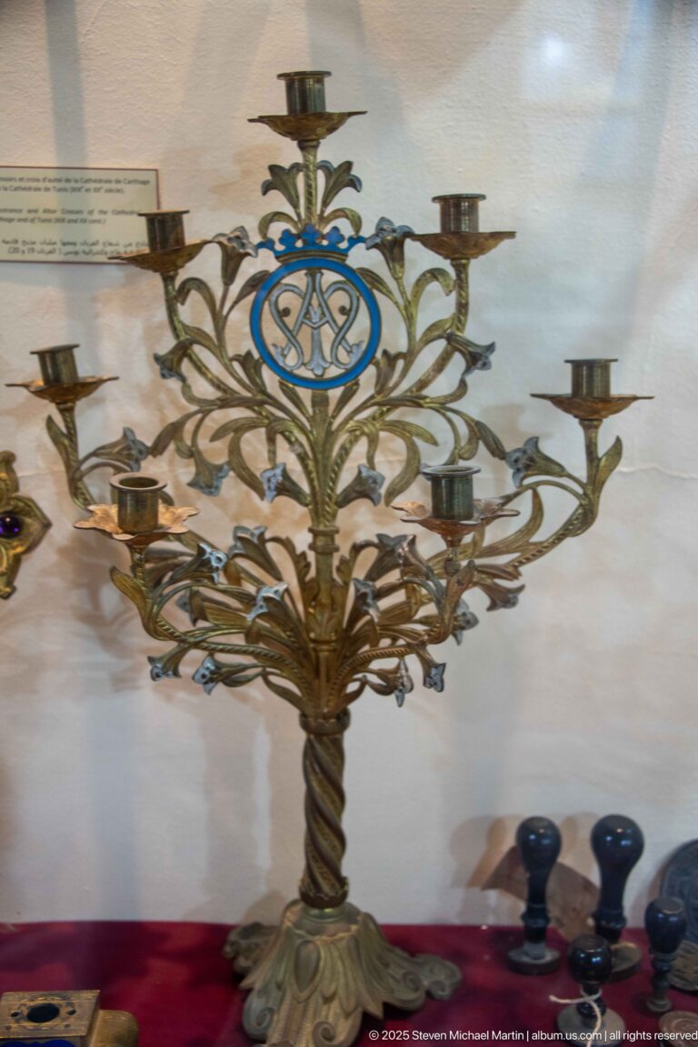 Branched metal candelabrum with multiple candle cups, curling foliage, twisted stem, and blue Marian monogram medallion.