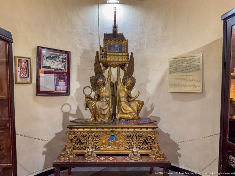 Gilded shrine-like devotional object with pinnacled canopy, two angelic figures, pierced base, and small colored details.