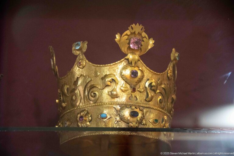 Heavy gilded crown with raised scrollwork, colored cabochon stones, curved points, and a floral crest above the central jewel.