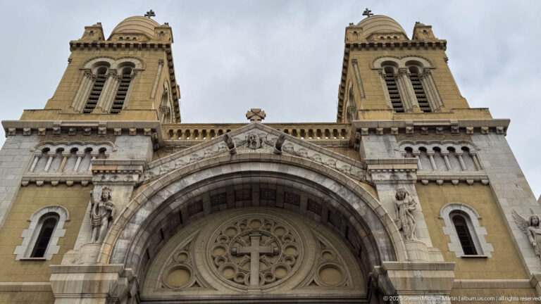 Upward view between the cathedral’s twin towers, with central gable, cross, statues, monumental arch, and circular tracery.