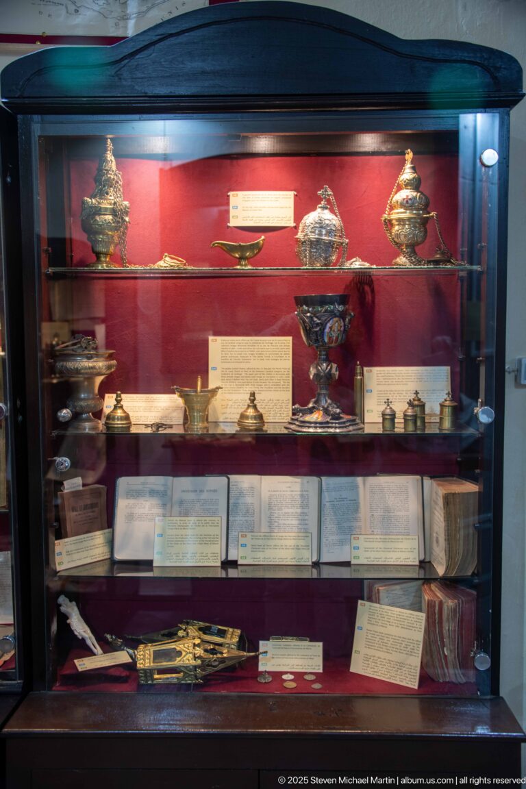 Large red-backed display case with liturgical vessels, ornate containers, printed documents, books, labels, and smaller ecclesiastical objects.
