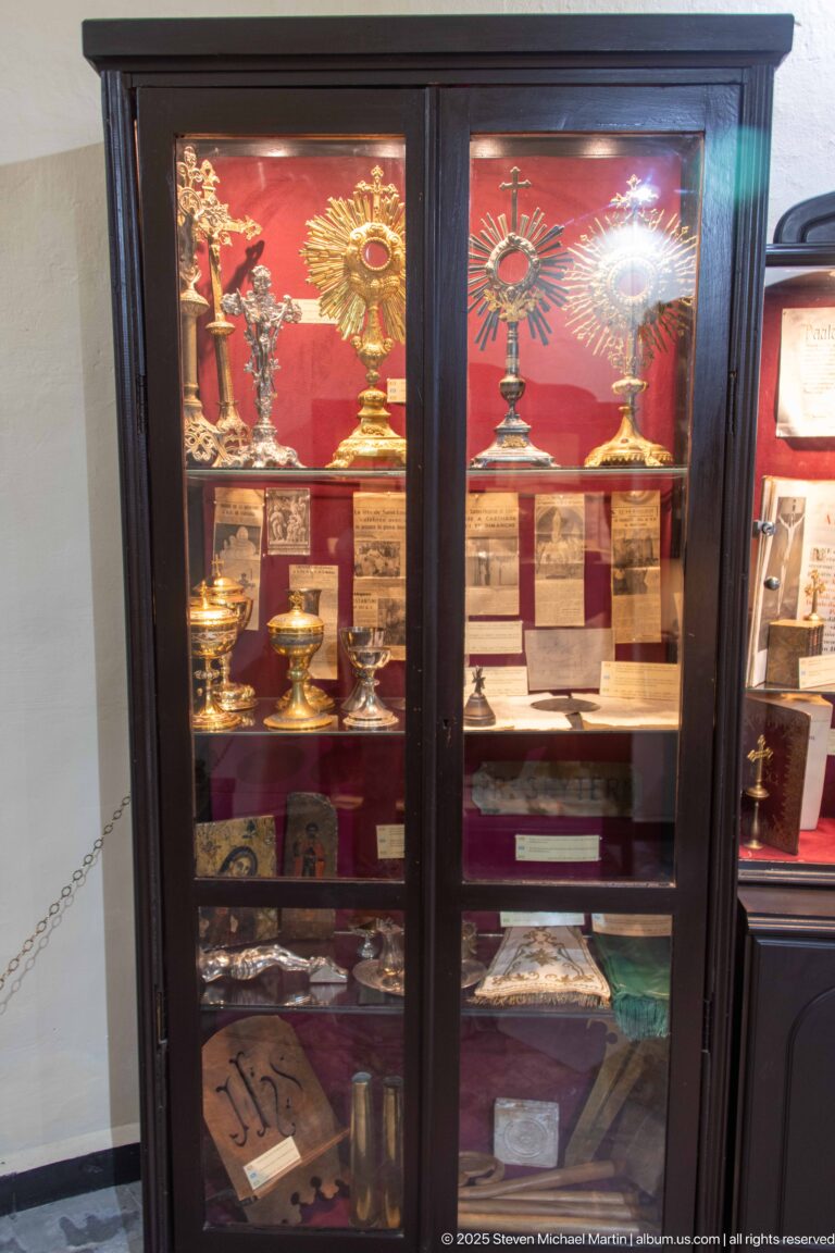 Tall glass cabinet with sunburst monstrances, metal vessels, books, documents, labels, and smaller devotional objects on red backing.