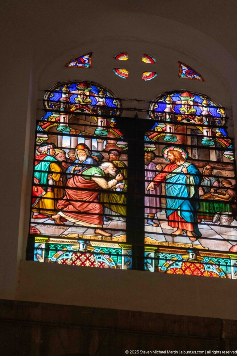 Paired stained-glass narrative scenes with grouped figures in red, blue, green, and gold beneath decorative floral roundels.