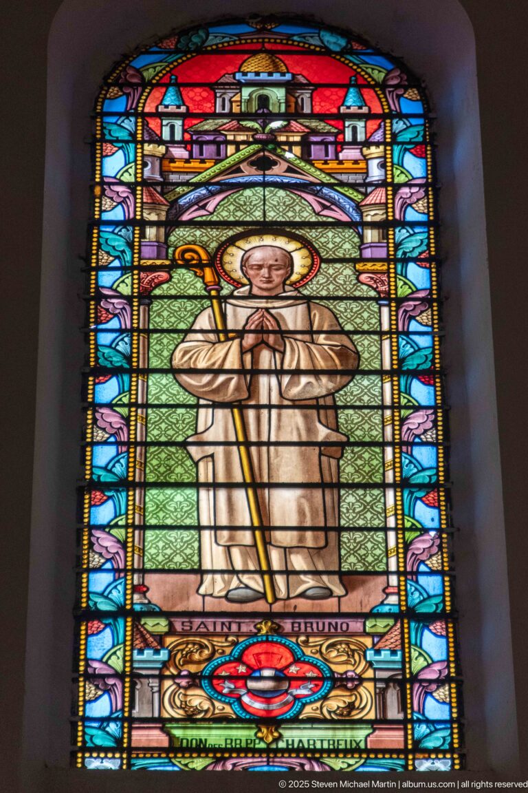 Stained-glass haloed male saint in a pale robe holding a long staff within an ornate multicolored architectural frame.