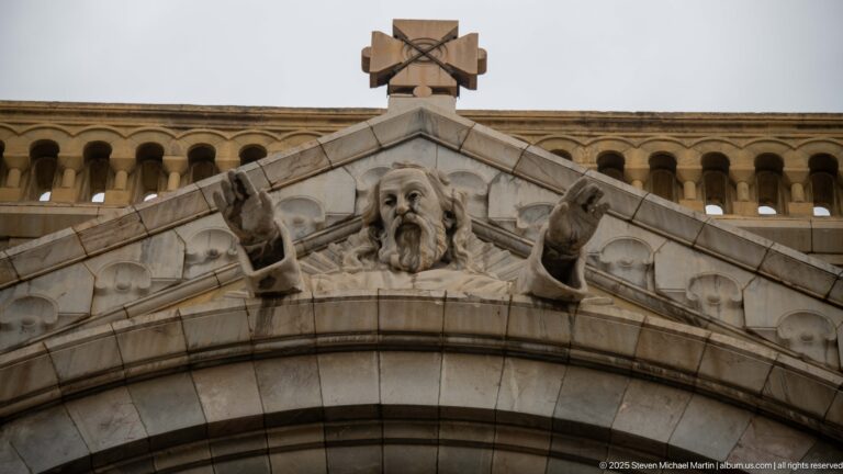 Bearded sculpted figure with raised hands beneath a stone cross at the apex of the cathedral entrance gable.