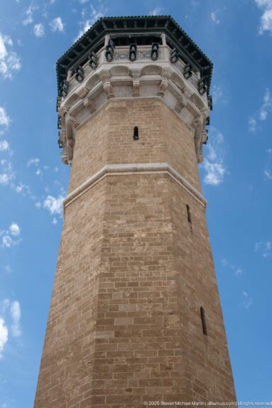 Low-angle view of an octagonal brick tower with slit openings, pale projecting upper stage, and dark roof.