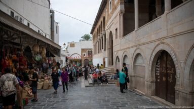 Busy medina lane with market stalls and pedestrians beside a long arcaded stone façade at the Zitouna Mosque precinct.